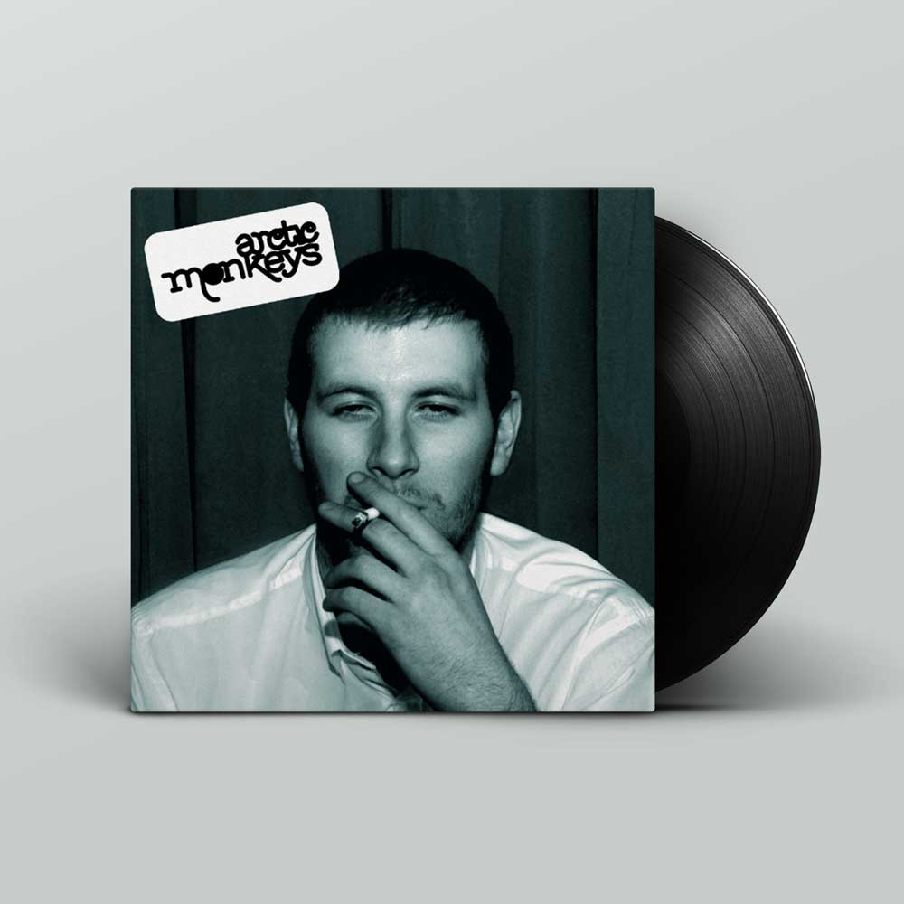 Arctic Monkeys - Whatever People Say I Am, That's What I Am Not (Vinyl LP). A vinyl record partially pulled from its album sleeve, shown on a light grey background. The sleeve features a black-and-white portrait of a man smoking, with a white sticker reading 'The Arctic Monkeys' in the top left.