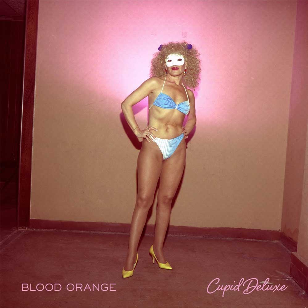 Blood Orange - Cupid Deluxe (Vinyl 2LP). A person stands in a room wearing a light blue and white bikini, a white eye mask, a curly wig, and yellow heels against a pink-lit wall. Text at the bottom reads 'BLOOD ORANGE' and 'Cupid Deluxe'.