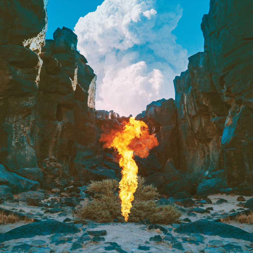 Bonobo - Migration (Vinyl LP 180g) record cover showing a flame rising from a rocky landscape under a blue sky with clouds.