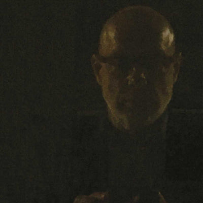 Brian Eno - Reflection (Vinyl 2LP) A very dark, low-key close-up of a bald man's face in shadow, with only faint warm highlights on his forehead and cheek against a black background.