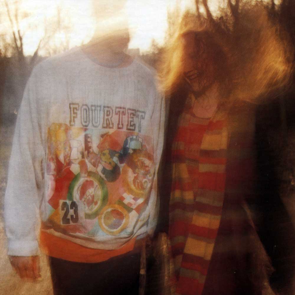 Four Tet - Pause (Vinyl LP 180g) A person wearing a light grey sweatshirt with a colourful retro graphic and the word 'FOURTET' on the front, standing outdoors in warm, backlit light next to another person.