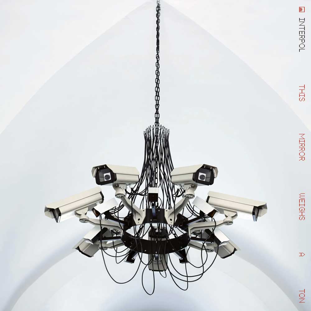 Interpol - This Mirror Weighs a Ton (Vinyl 2LP Transparent Red) Vinyl record album cover showing a multi-camera ceiling chandelier featuring numerous white security cameras with black lenses and numerous black cables, suspended from a black chain against a white arched ceiling.