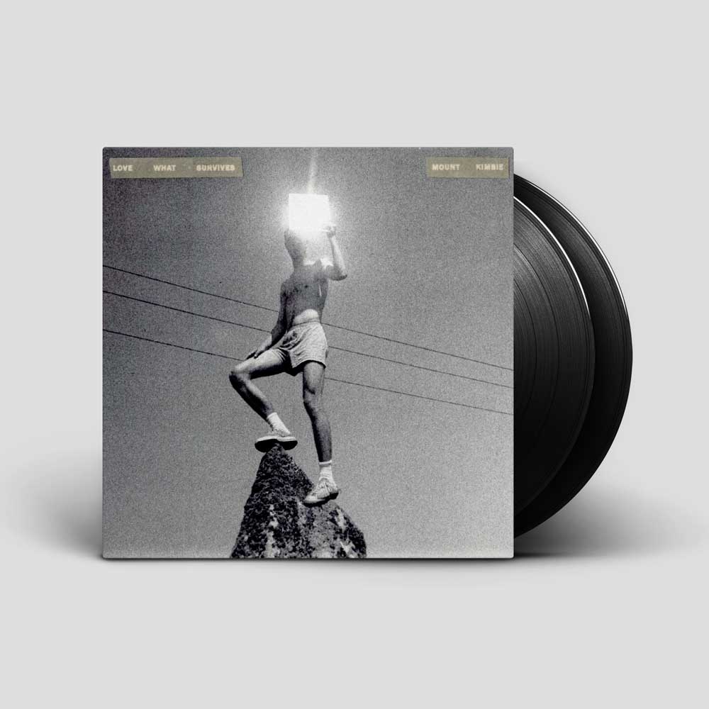 Mount Kimbie - Love What Survives (Vinyl 2LP) A vinyl record album shown standing upright with part of the black record visible behind the cover. The cover art is a black-and-white photo of a person standing on a rock holding a glowing light source, with small text at the top reading 'LATE WHAT HAPPENS'.