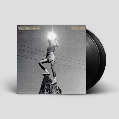 Mount Kimbie - Love What Survives (Vinyl 2LP) A vinyl record album shown standing upright with part of the black record visible behind the cover. The cover art is a black-and-white photo of a person standing on a rock holding a glowing light source, with small text at the top reading 'LATE WHAT HAPPENS'.
