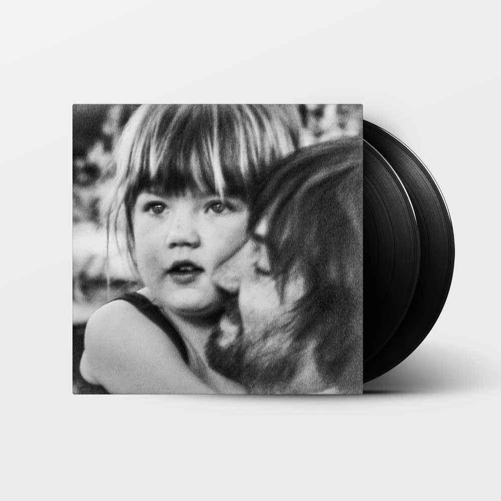 Tame Impala - Deadbeat (Vinyl 2LP) A vinyl record partially visible behind its square album cover, which features a black-and-white photograph of a young child looking at the camera while being kissed on the cheek by an adult. The record itself is black, set against a clean light gray background.